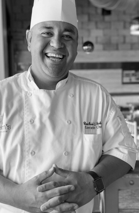 Executive Chef Rachid Hamdane stands proudly inside Kareem's Lebanese Kitchen, wearing a traditional chef's coat and hat, welcoming guests with a warm smile.