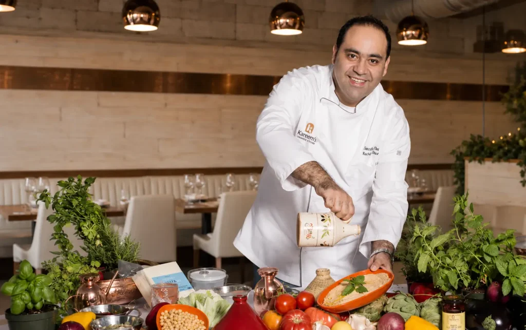 Executive Chef Rachid Hamdane prepares a traditional Lebanese dish inside Kareem's Lebanese Kitchen, surrounded by fresh herbs, vegetables, and authentic ingredients in a warm, inviting restaurant setting.