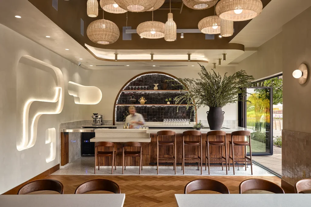 Gaithersburg bar with an arched dark-tile shelf, woven rattan pendant lights, walnut stools and glass doors open outside