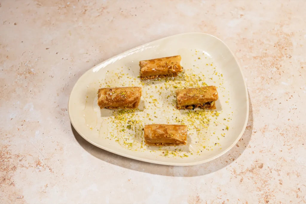 Four rolled baklava fingers glazed with syrup on a cream plate sprinkled with crushed pistachio