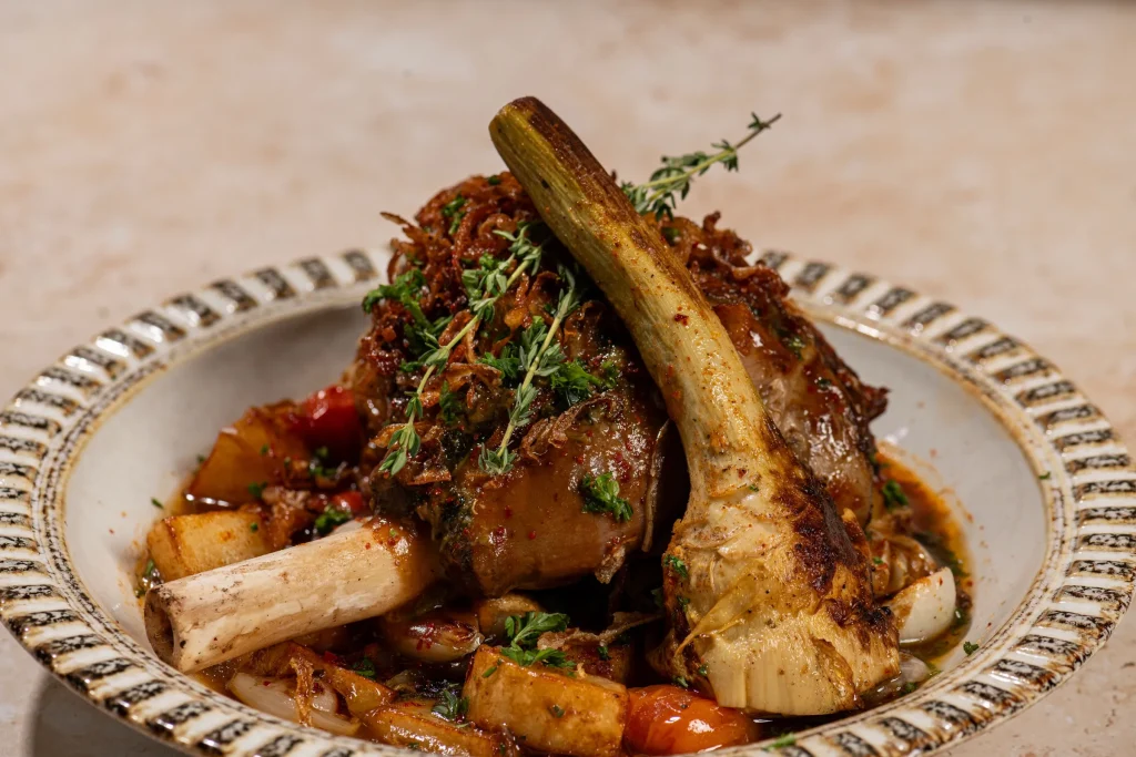 Braised lamb shanks with crispy onions and thyme over roasted potatoes, tomatoes and onions in a patterned bowl