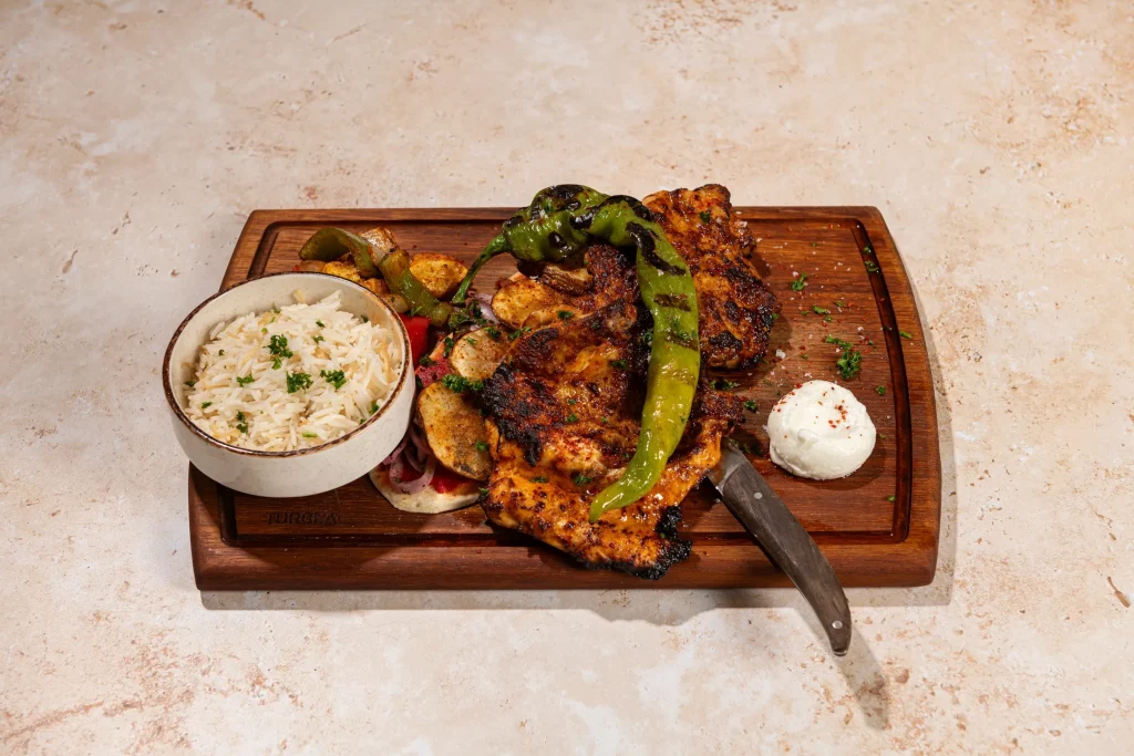 Charred grilled chicken with a blistered green pepper, potatoes, rice bowl and white dip on a wooden board with a knife