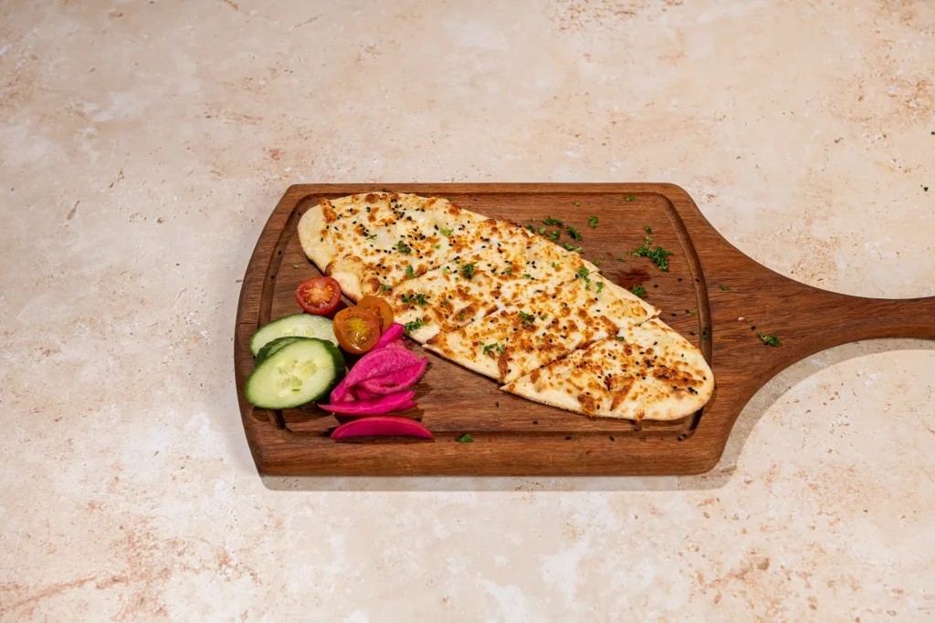 Sliced cheese flatbread with sesame and nigella seeds on a paddle board with cucumber, tomatoes and pickled turnips