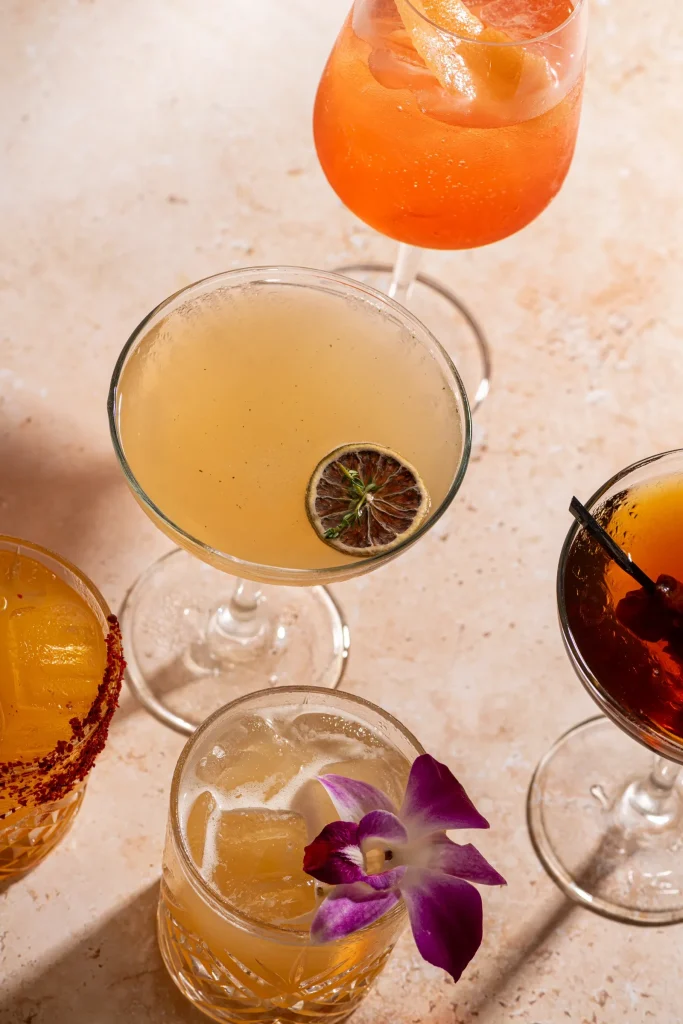 Overhead of five cocktails: spritz, coupe with dried lime, orchid-garnished drink, chili-rimmed glass and dark stirred drink
