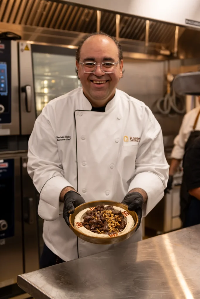 Chef Rachid Eido in a white jacket holding a bowl of hummus topped with beef and pine nuts in the kitchen