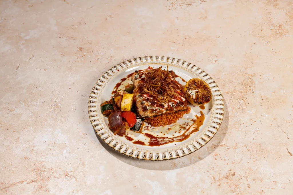 Fish fillet with crispy fried onions on red-tinted grain with roasted vegetables and charred lemon on a patterned plate