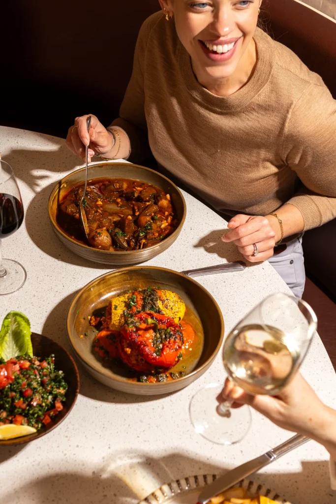 Smiling woman at a table with a mushroom stew, a roasted stuffed vegetable dish, tabbouleh and glasses of wine