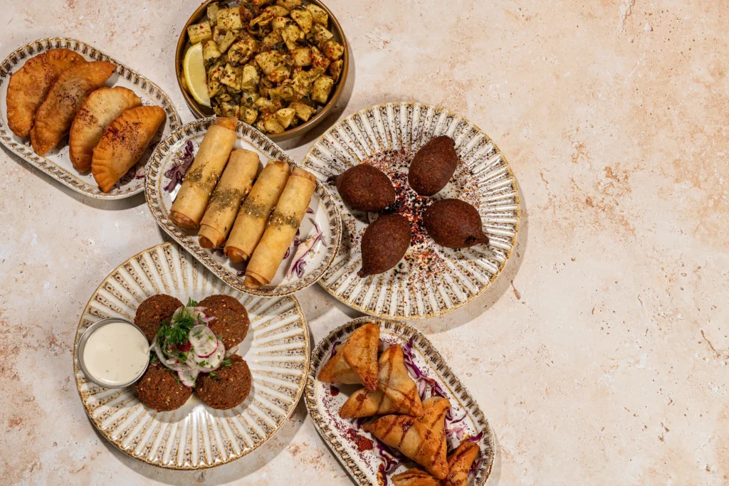Overhead of fried pastries, cheese rolls, kibbeh, falafel, grilled triangle pastries and spiced potatoes on patterned plates