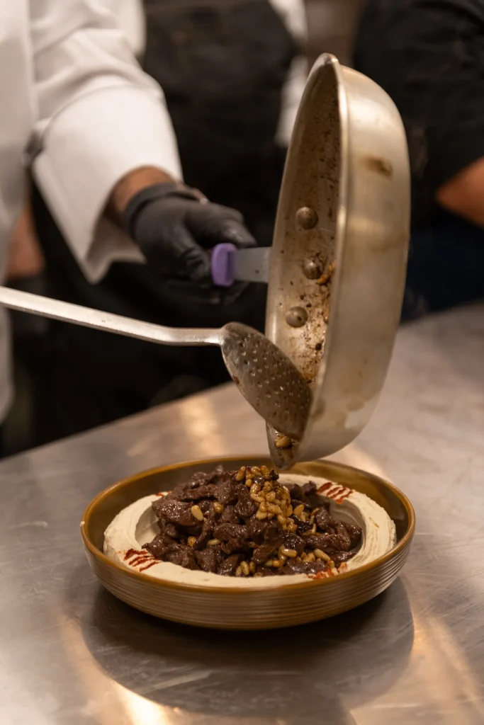 Gloved chef spooning sautéed beef and toasted pine nuts from a pan onto a bowl of hummus