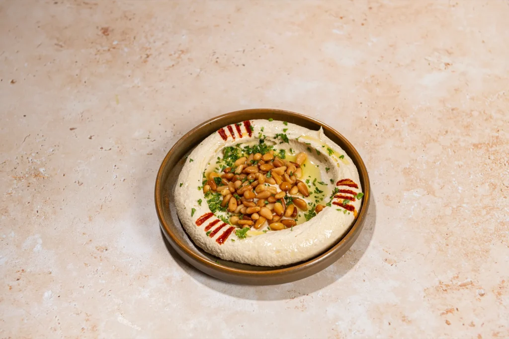 Hummus topped with toasted pine nuts, chopped parsley, olive oil and paprika stripes in a brown bowl