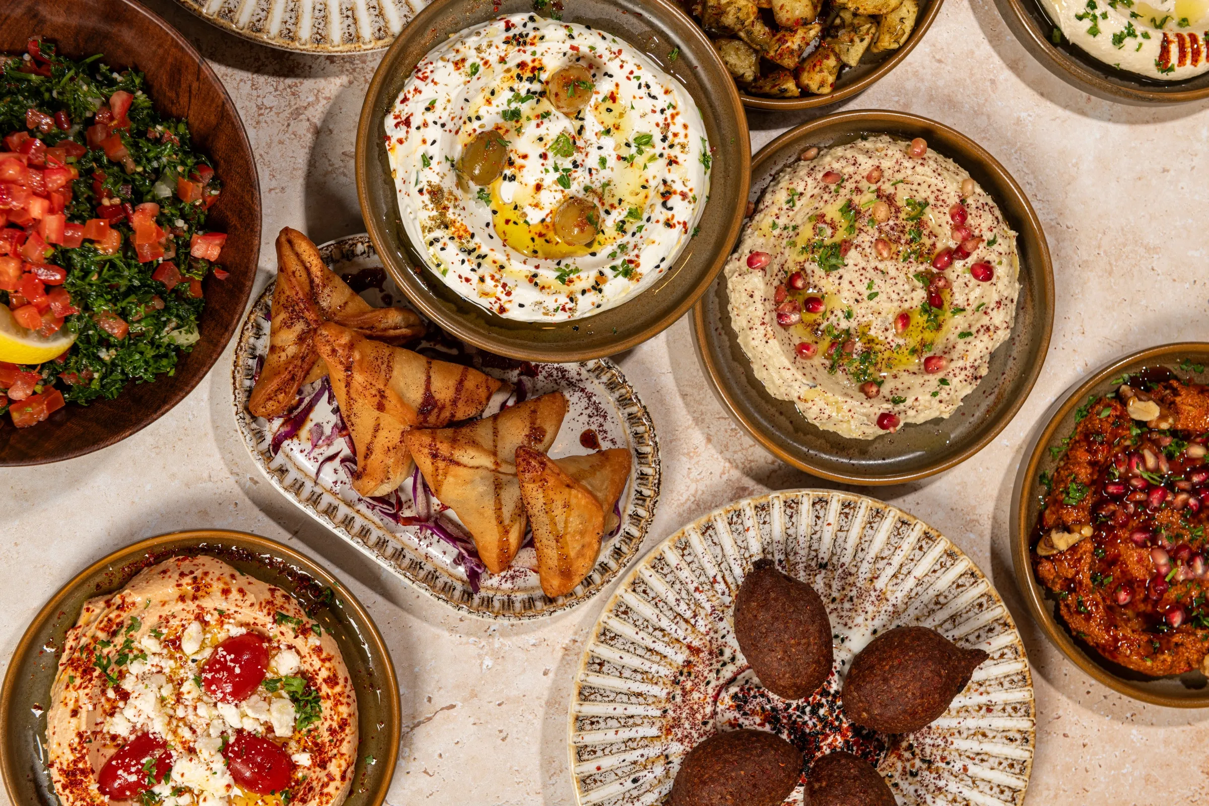 Overhead mezze of labneh, tabbouleh, grilled pastry triangles, kibbeh, muhammara and hummus with feta and tomatoes